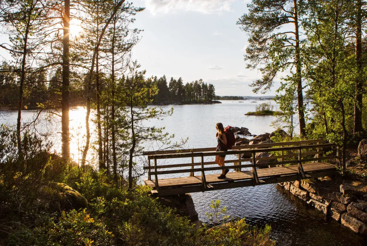 Vandrare korsar en bro i Glaskogens naturreservat. Solen skiner genom träden över en sjö.