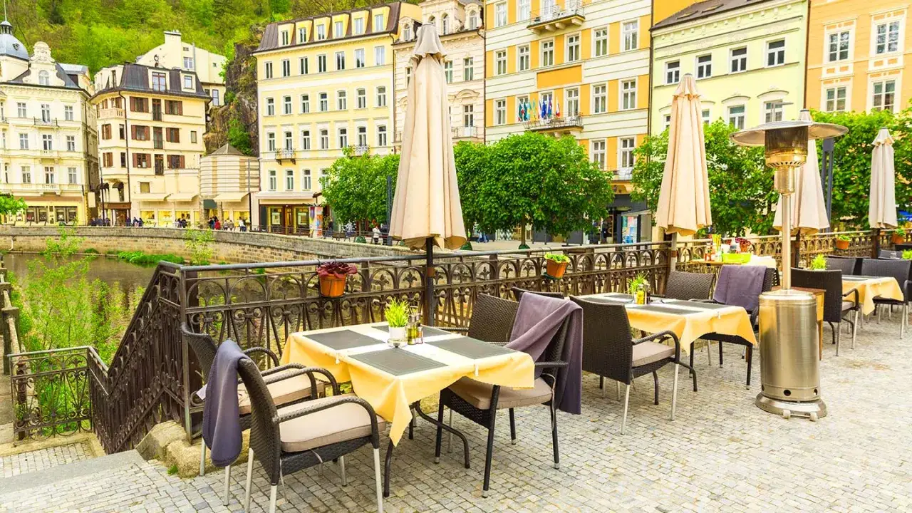 Uteservering vid en flod i Karlovy Vary med vackra byggnader i bakgrunden.