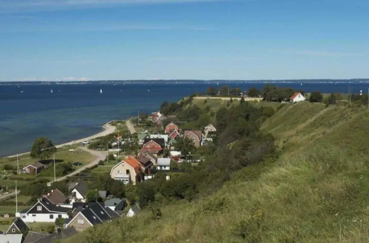 Vacker kustvy över små orter i Skåne med röda tak och gröna kullar mot ett blått hav med segelbåtar.