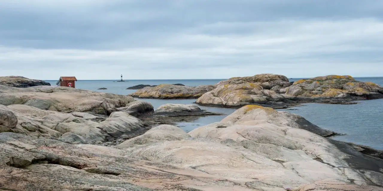 Klippigt landskap vid tjurpannans naturreservat med en liten röd stuga och ett fyr i fjärran.