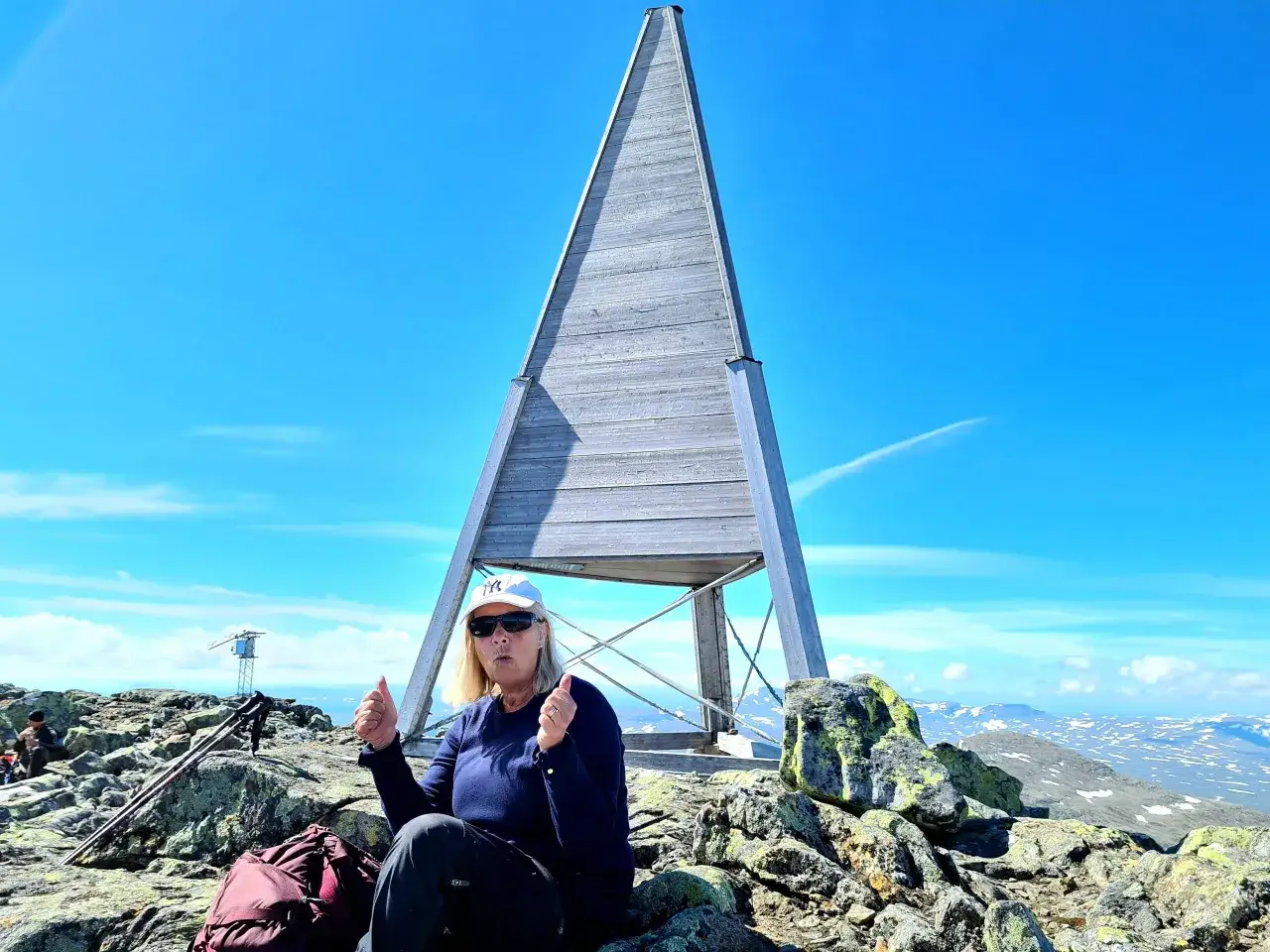 En kvinna firar sin helags topptur med tummen upp vid en pyramidliknande struktur på en stenig bergstopp under en klarblå himmel.