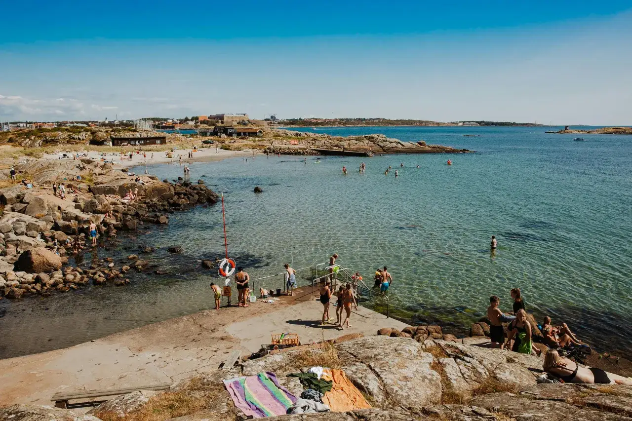 Människor njuter av en solig dag vid havet, ett perfekt utflyktsmål i Halland. Barn och vuxna badar och solar på klippor och strand.
