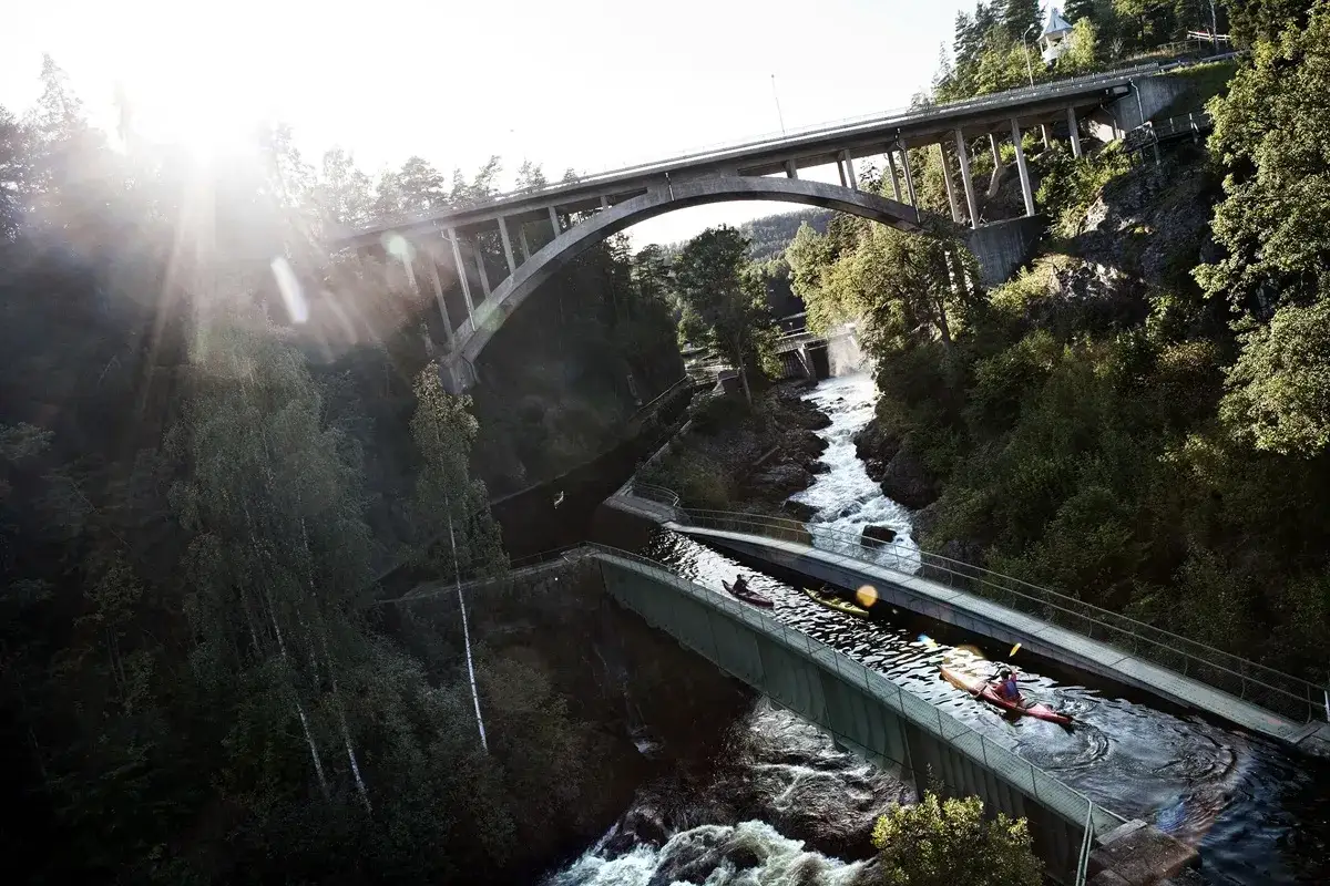 En person paddlar kajak i Dalslands kanal, omgiven av lummig grönska och dramatiska broar.