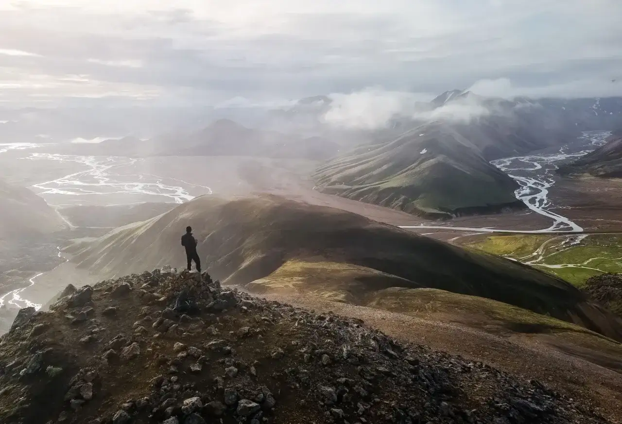 En person står på en klippig topp och blickar ut över ett dramatiskt landskap med slingrande floder och dimhöljda berg. En fantastisk vy för den som vill vandra på Island.