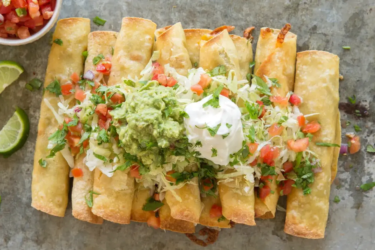 En tallrik med gyllene, krispiga taquitos, toppade med guacamole, gräddfil, sallad och tomater. Ett perfekt taquitos recept för alla tillfällen.