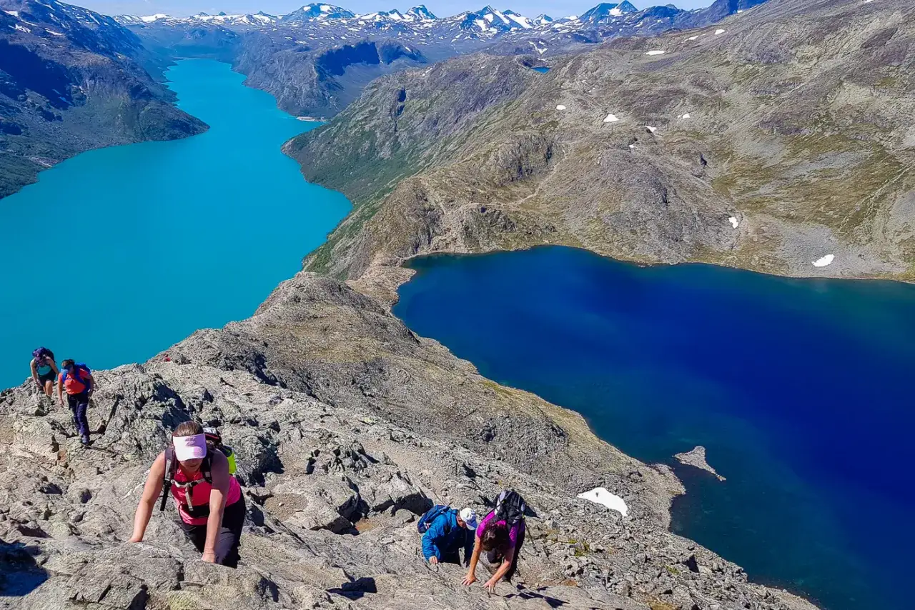 Vandrare klättrar på en stenig stig med turkosa och blå sjöar nedanför. En fantastisk vy som påminner om en Besseggen vandring.