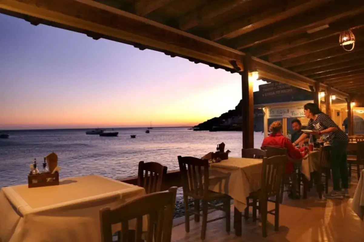 En kväll vid havet under en resa till Grekland. En taverna med dukade bord och gäster som äter medan solen går ner över vattnet.