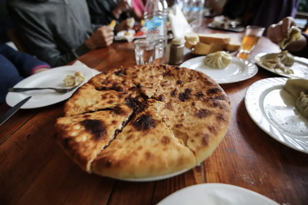 En stor, gyllene khachapuri, en klassiker inom georgisk mat, delas upp på ett träbord. Runtom syns khinkali och andra läckerheter.