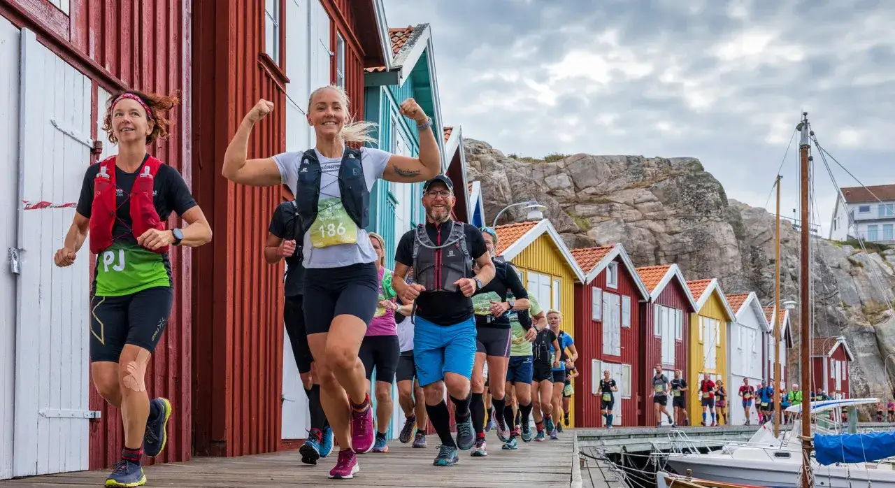 Löpare i en tävling längs en träbrygga med färgglada hus i bakgrunden. En kvinna visar muskler, en härlig icebug experience.