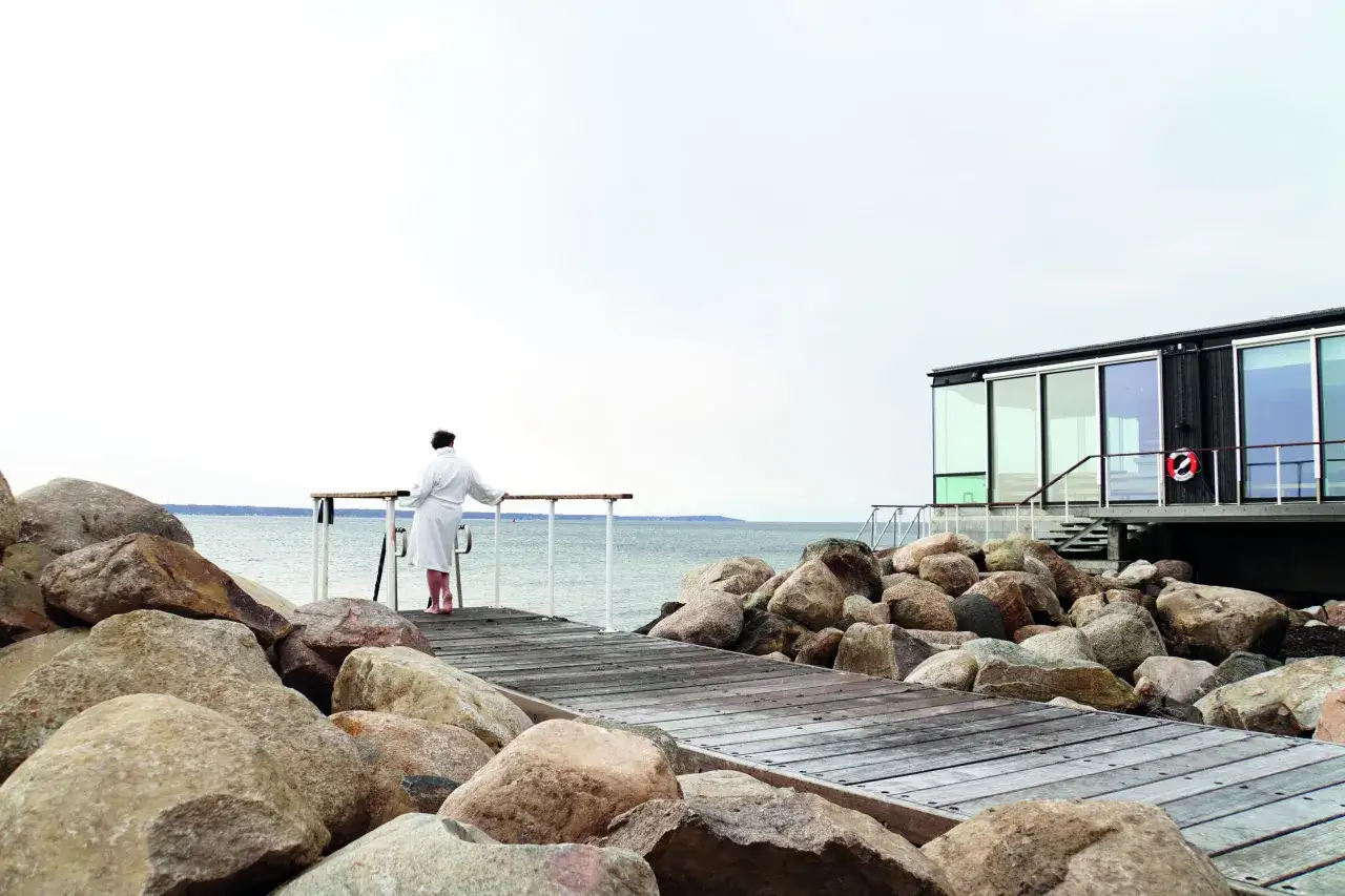 En person i morgonrock njuter av utsikten vid ett utomhusbad i Skåne. En modern byggnad med stora fönster syns i bakgrunden.