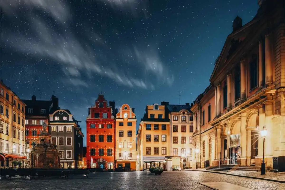 Färgglada hus i Gamla stan under en stjärnklar natt. En stenlagd plats med en fontän i förgrunden.