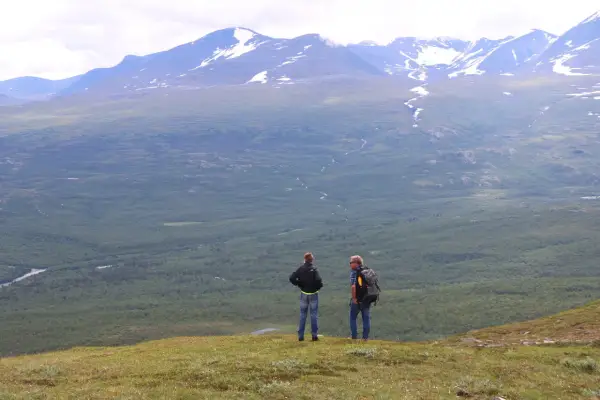 Två vandrare njuter av utsikten över vidsträckta fjäll och skogar, kanske under en vandra abisko 3 dagar. Snöklädda berg tornar upp sig i bakgrunden.