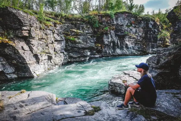 En person sitter vid en forsande turkos flod omgiven av klippor. Ett perfekt utflyktsmål i Mellansverige för naturupplevelser.