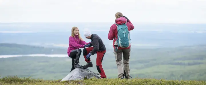 Två barn hjälper varandra upp på en sten under en vandring. En vuxen står bakom med ryggsäcken redo för äventyr.