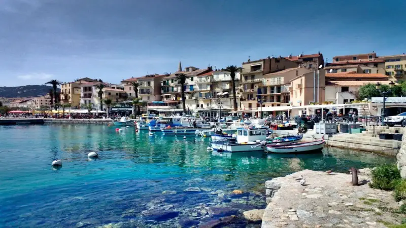 Båtar i en turkos hamn i Calvi, Korsika, med färgglada hus och palmer längs kajen.
